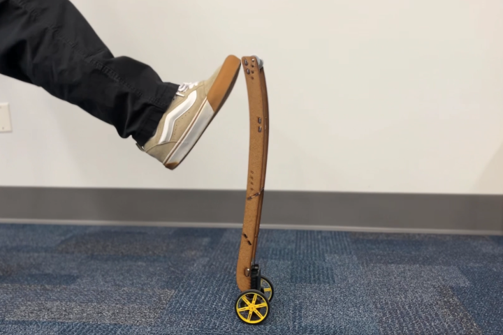 A student's foot is shown trying to tip over an inverted pendulum, made out of wood and with yellow and black wheels at bottom.