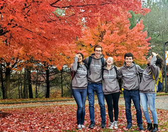 Five students smiling or laughing while wearing matching Olin sweatshirts and standing in front of bright orange and red trees in autumn, with fallen leaves on the ground