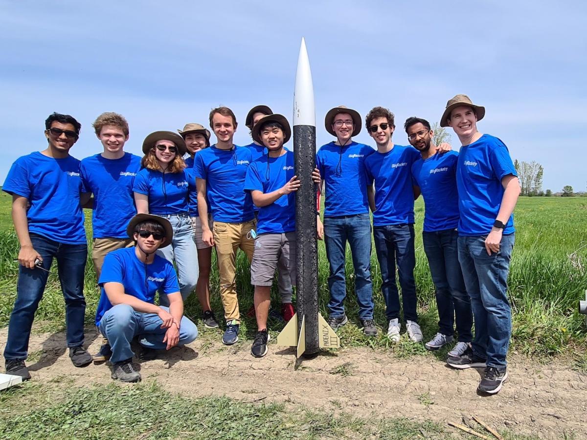 Members of the Olin College Rocketry Team in blue shirts pose in front of one of their rockets.