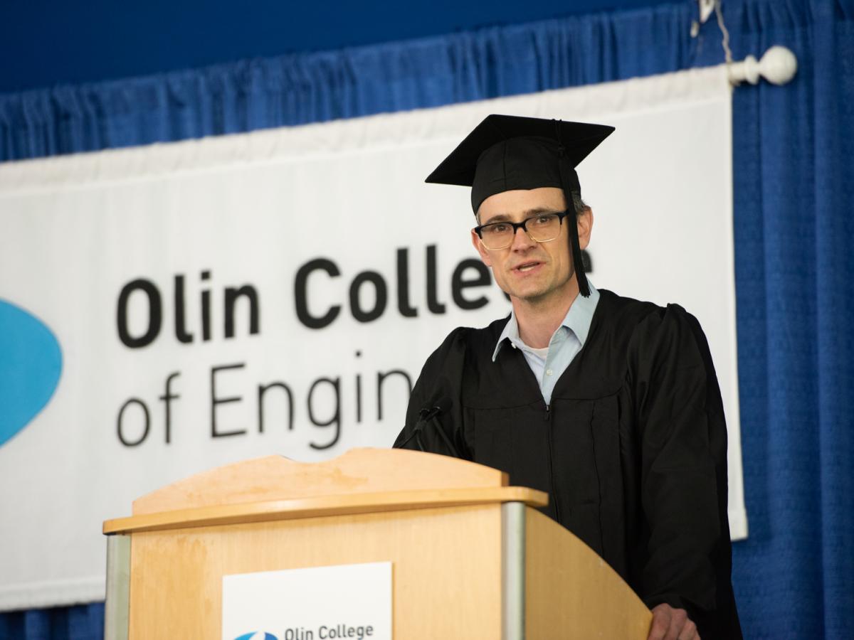 Tim Sauder, Associate Professor of Practice in Design, in a black cap and gown, stands at a podium with the Olin College logo behind him. 