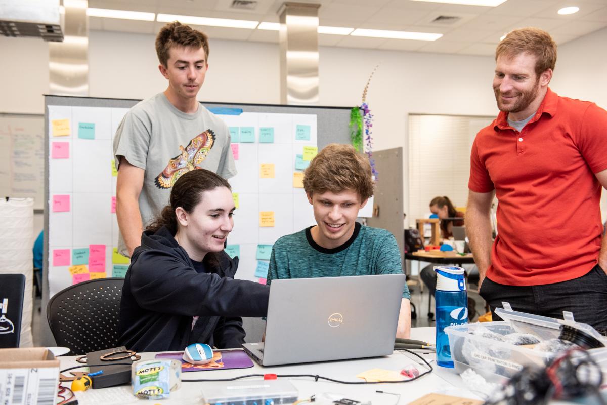 Two sitting students and one standing student in grey shirt confer with a standing professor in orange shirt over a macbook laptop and some new research in a classroom.