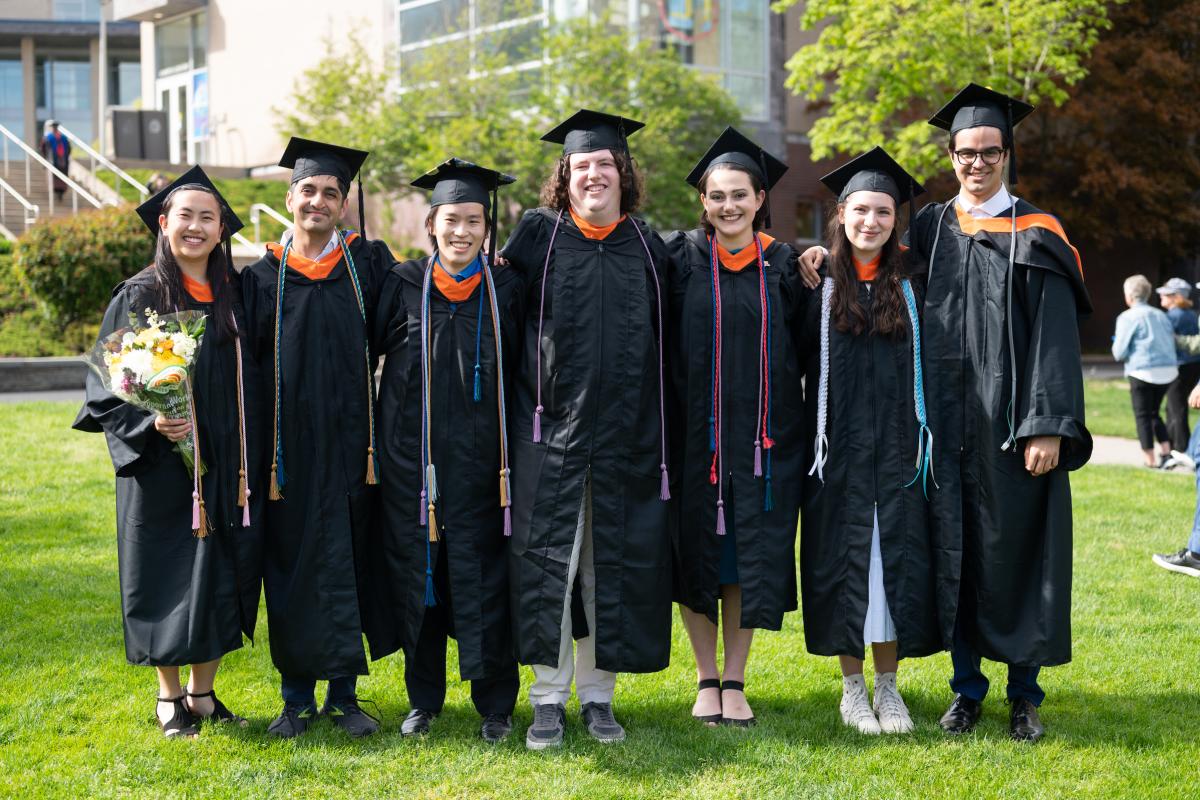 Seven Olin College of Engineering graduates pose in a line with their cap and gowns on. 