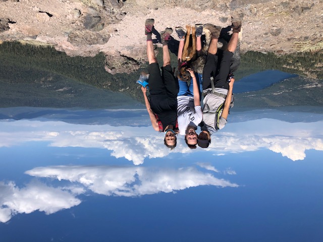 Three people and a dog hiking in front of a scenic overlook.