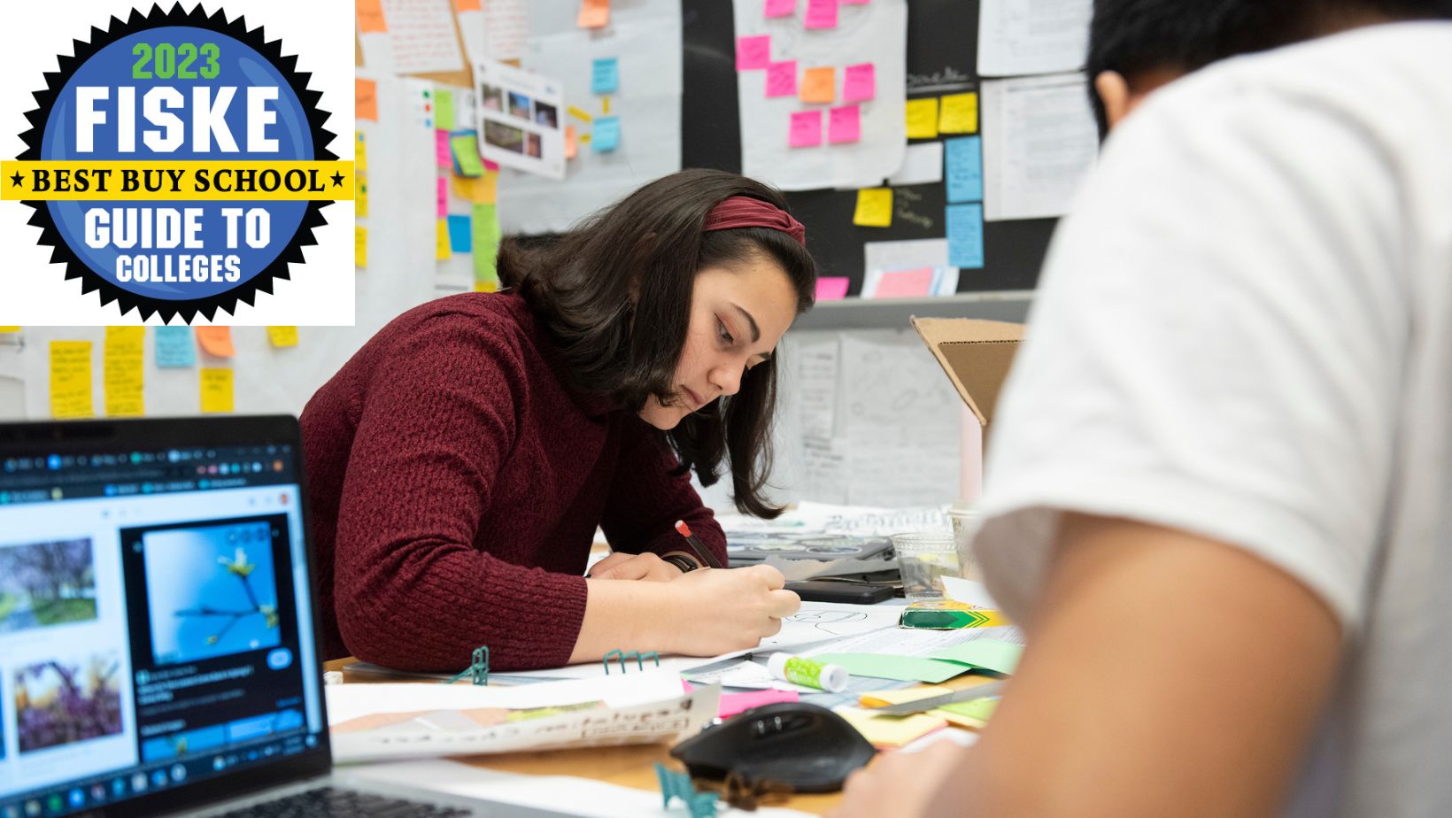 An Oliner is writing on a table in a sticky-note filled classroom.