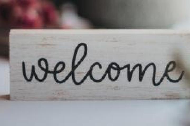 A small sign that says "welcome" in front of an out-of-focus teapot and other items on a table.