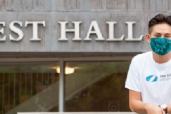 A person standing outside a building labeled "West Hall," wearing an Olin t-shirt and a mask.