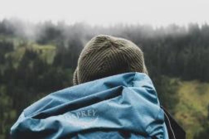 A backpacker shown from the back with a blue backpack and a green beanie, hiking through a green, hilly area in light fog.