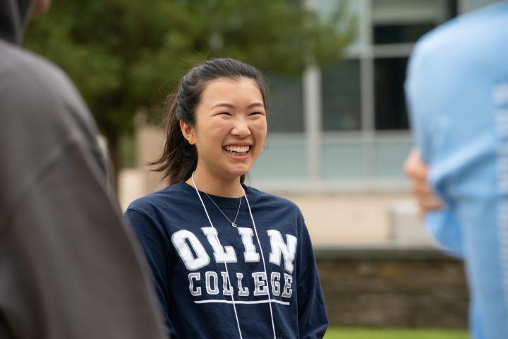 A dark haired student with a navy "Olin College" sweatshirt smiles candidly for the camera while on Olin's campus.