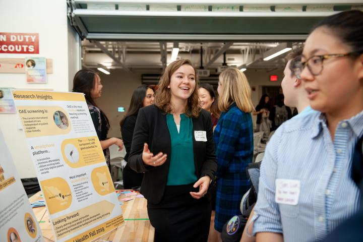 A student in green blouse and black blazer talks with guests in front of a poster board.