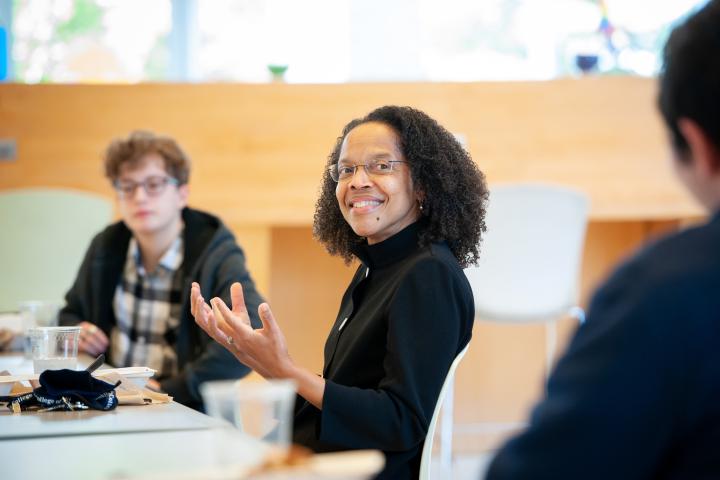 Olin President Gilda Barabino in black shirt and glasses sits with students in the Dining Hall during lunch.