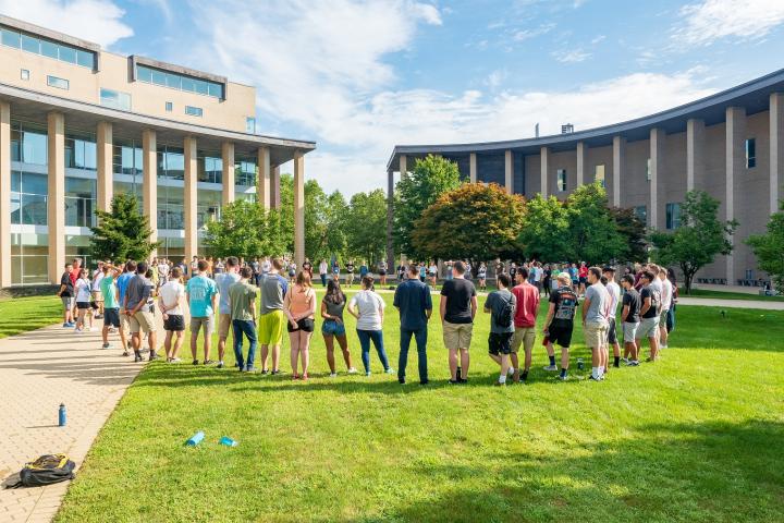 Olin students in a circle on the oval under a blue sky.