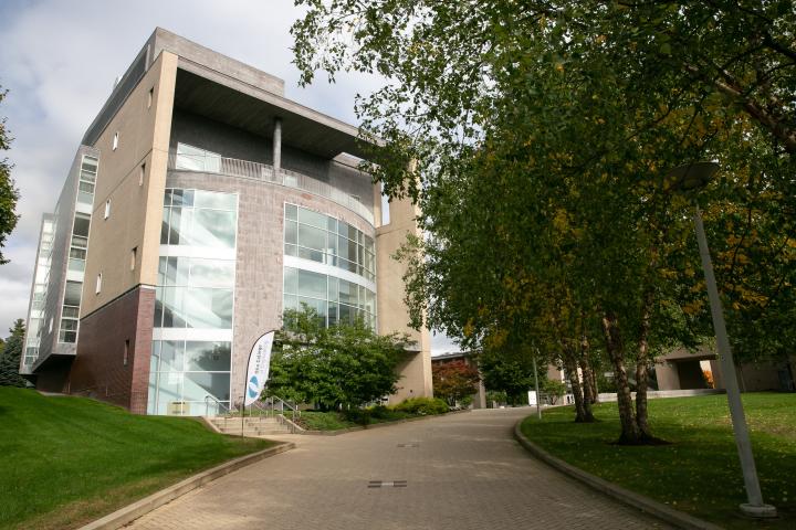 Exterior view of a building with windows running up it and trees obscuring what's next to it.