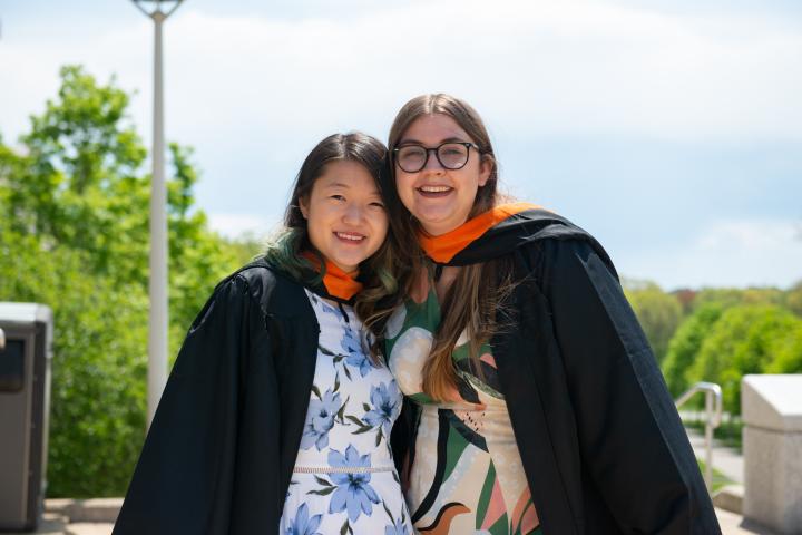 A photo of two graduates smiling with blue sky and green trees in the background