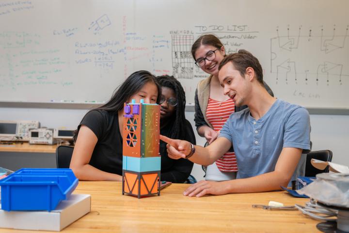 Four students work together in a classroom, pointing to a multicolored Cube prototype.
