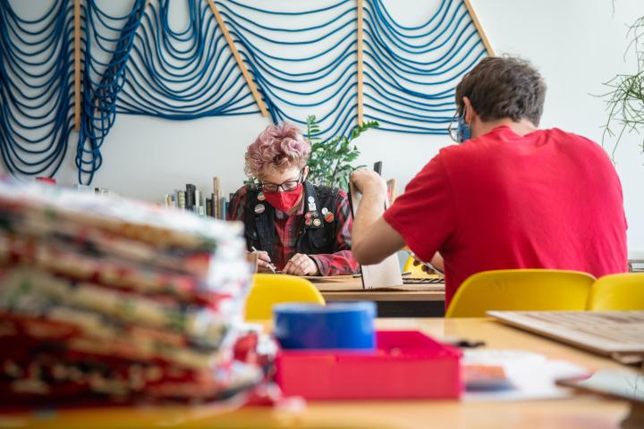 Two students work at a table, with multi-colored cloth in the foreground and a decorative rope wall hanging in background.