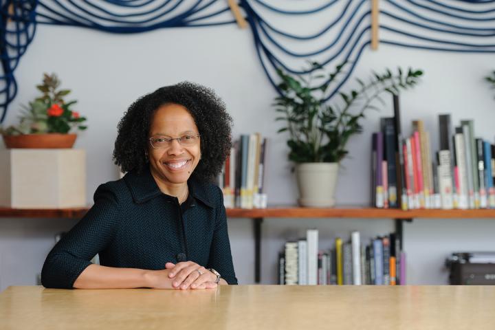 Olin College President Gilda A. Barabino, in a dark top and glasses, sits at a wooden table and smiles for the camera.