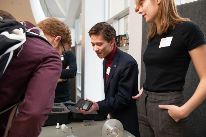 Two students - both in black tops, stand behind a table and show another person in maroon shirt and backpack a smartphone screen.