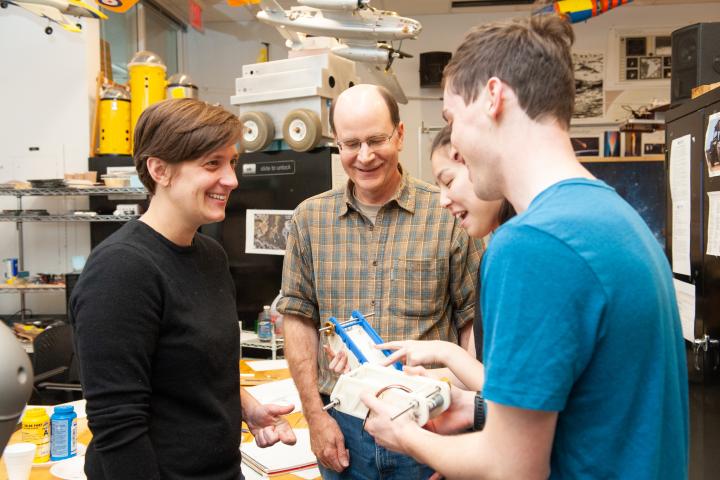 Professor Dave Barrett (in the center) with checkered shirt and glasses, and Professor Daniella Faas (on the left) in black shirt and close-cropped hair, talk with students in a machine lab in 2019.