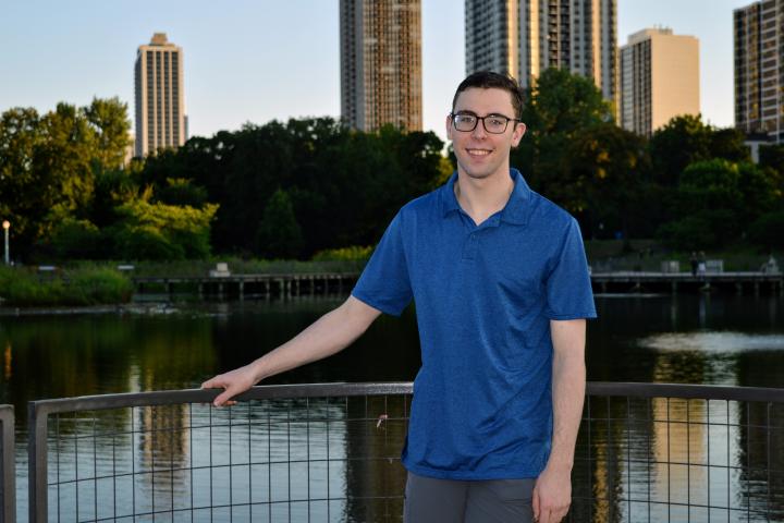 A photo of a young man standing near a fence with a city skyline in the background