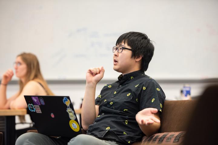 A man with glasses and dark hair, seated in a classroom with a laptop resting on his lap, talks to the class. 