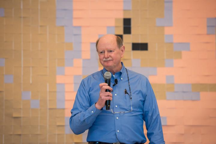 Olin's first president Rick Miller in blue dress shirt holds a mic in front of a wall of peach sticky notes.