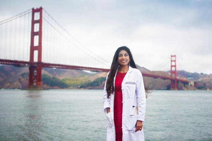A women in white lab coat and red outfit underneath, poses in front of the Golden Gate Bridge.