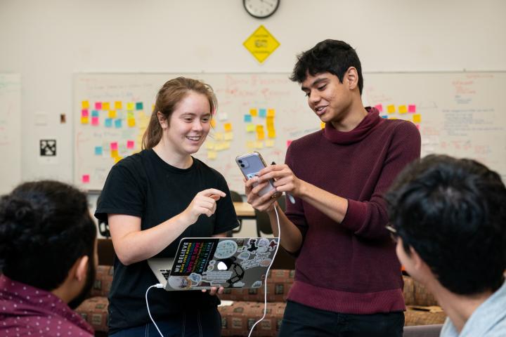 Two summer research students in dark clothing connect a phone and app to a laptop via a white cord as two other team members look on.