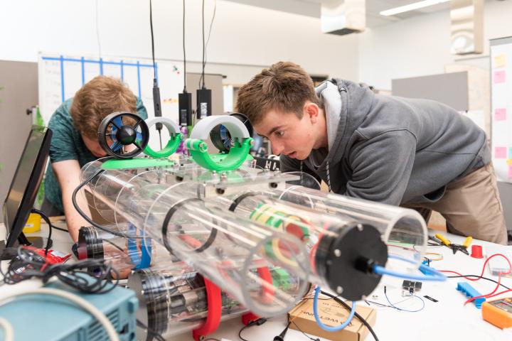 Two students on the Summer Research team lean over and work on making adjustments to three clear plastic cylinders.