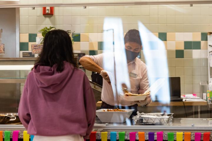 A student waits for a plate of food during a special Convocation Lunch in 2021. A person in a light purple sweatshirt waits for a masked employee to serve a plate of food.