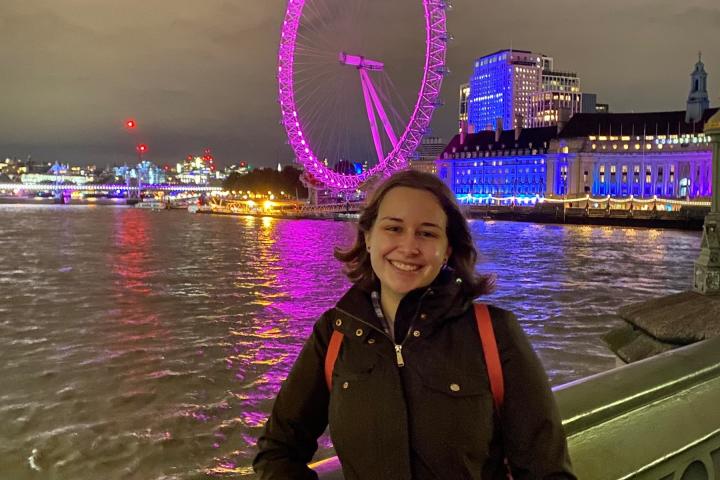 A young student smiles and poses for a photo on the waterfront with a florecent pink observation wheel in the background.