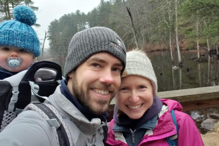 Leif hiking Great Brooks Farm with family. A man with a grey knit cap, brown facial hair and cute baby on his back, poses outside with a smiling woman in a light colored taupe knit cap and pink coat.