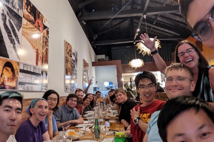 A large group of people take a selfie while sitting and standing at a long table filled with dinner glasses and plates.