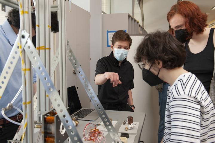 Braden Oh '23 (center), points to the thruster while interacting with students and members of the Olin community during Spring Expo 2022. A student wearing a blue face mask points to something on a table, with a large silver apparatus in the foreground.