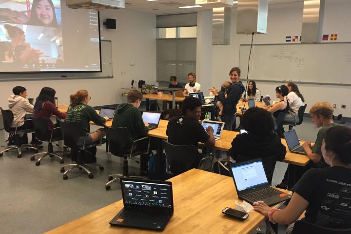 A women holding a baby stands in the middle of a classroom with students around her working on laptops