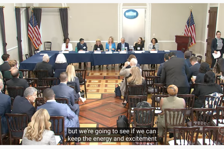 A panel of seven speakers sit in front of a crowd at a long table at the Whitehouse.
