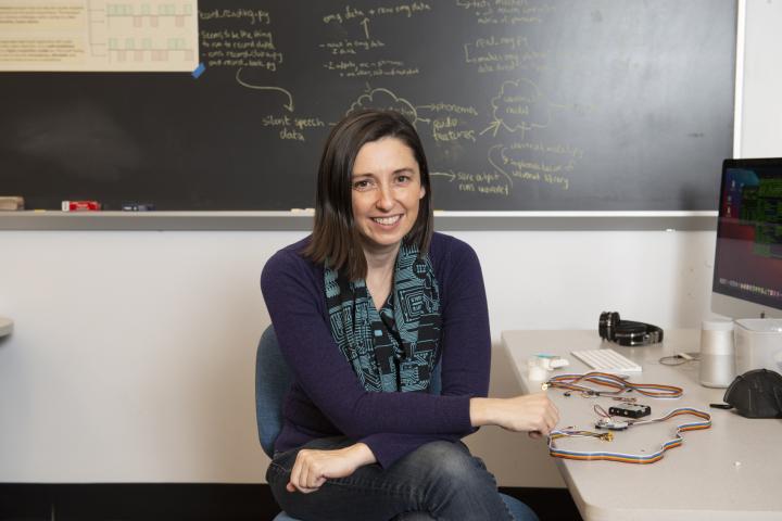 Woman with medium length brown hair and purple shirt, Professor Sam Michalka, sitting on desk.