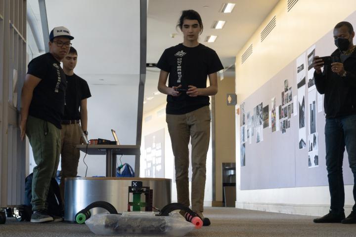Ethan '25, William '25, and Devlin '25 with their PIE final project, "HOVERBOIS." Four students stand around a robotic device on the floor, with one of them filming it with his smartphone.