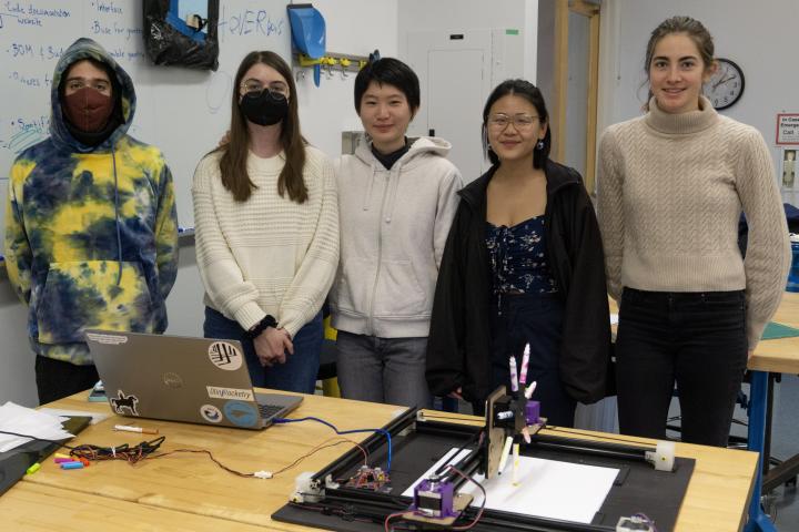 Five students stand in a row in front of a brown classroom table that has a laptop and a rectangle device on it. Five students stand in a row in front of a brown classroom table that has a laptop and a rectangle device on it.