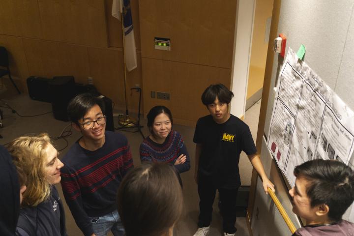 A photo taken from above five Olin College students as a couple of them look upward toward the camera. A photo taken from above five students as a couple of them look upward toward the camera.