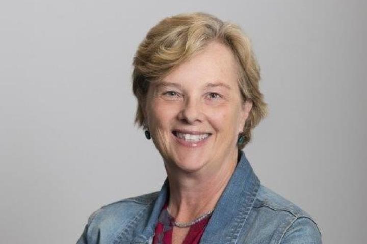 Susan Brisson Woman with short blonde hair stands, smiling with arms crossed, wearing a jean jacket and red shirt.