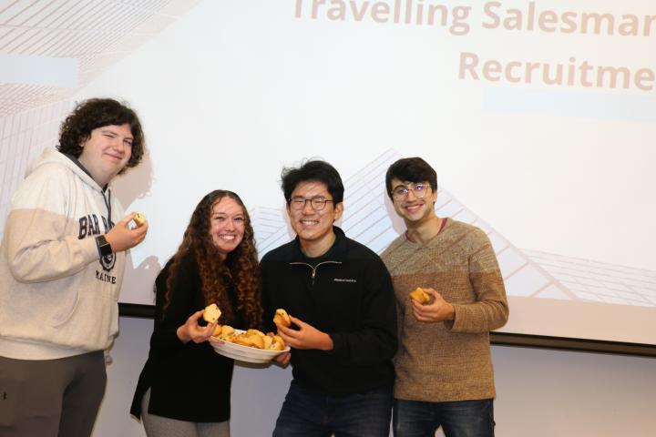 Four students stand in front of a presentation screen, holding muffins.
