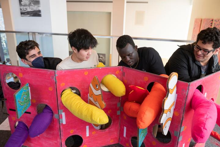 Four students crouch down behind a red cardboard structure while wearing bright colored hand coverings. They are sticking their covered hands through the holes in cardboard.