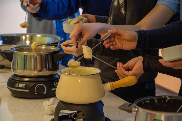 A photo of three sets of hands dipping pieces of food on metal sticks into cheese fondue.