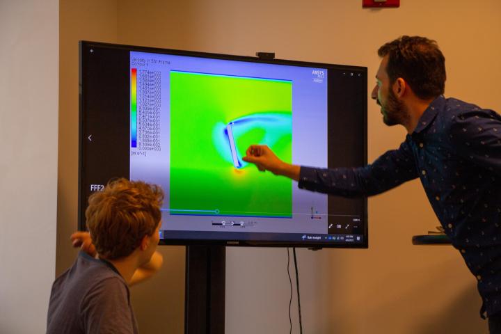 A man points to a glowing green computer screen while a student looks on. 