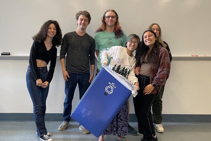 Six students pose for a group photo with a blue recycling bin.