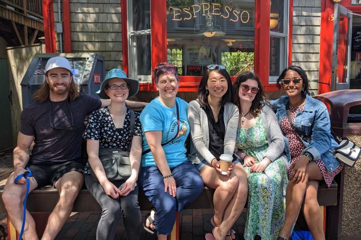 Six smiling friends pose for a group photo outdoors on a bench in front of a building that says, "Espresso."