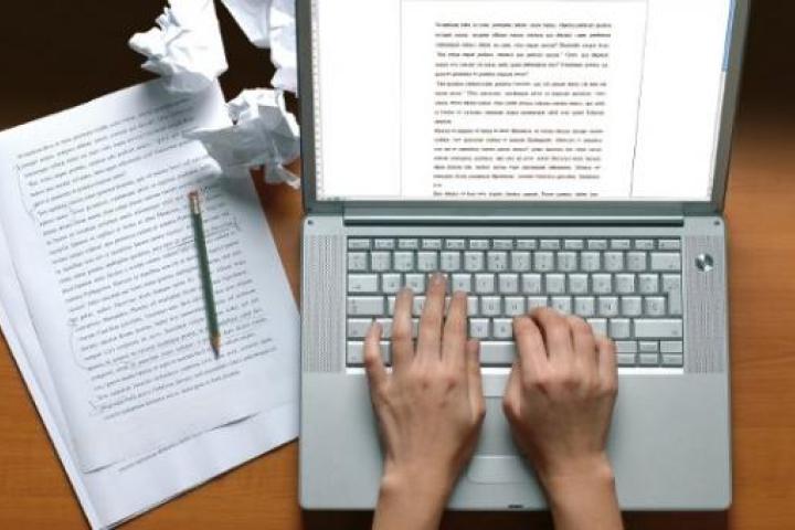 Person typing an essay on a laptop, viewed from above. Scrap pieces of paper with writing on the left side of the laptop and crumbled up balls of paper.