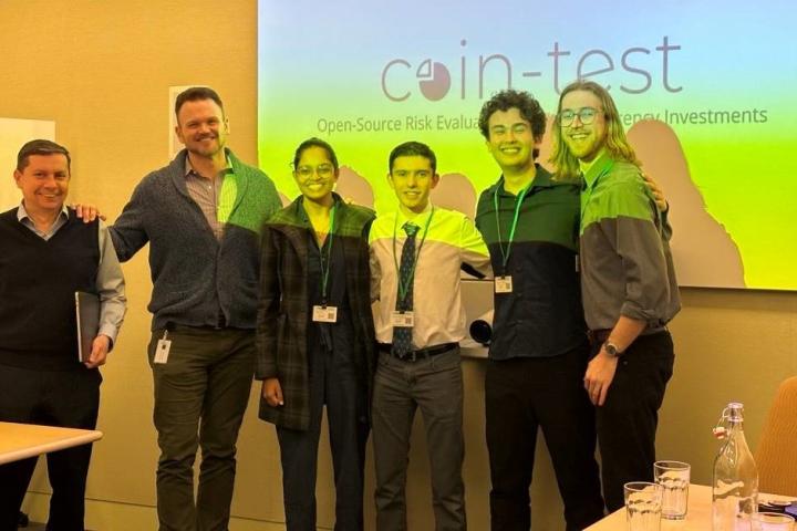 Four students pose with two employers in front of a large presentation screen, bathing the group in hues of orange and yellow.