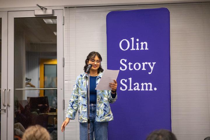 An Olin student is pictured reading from a piece of paper while standing in front of a purple flag that reads "Olin Story Slam."