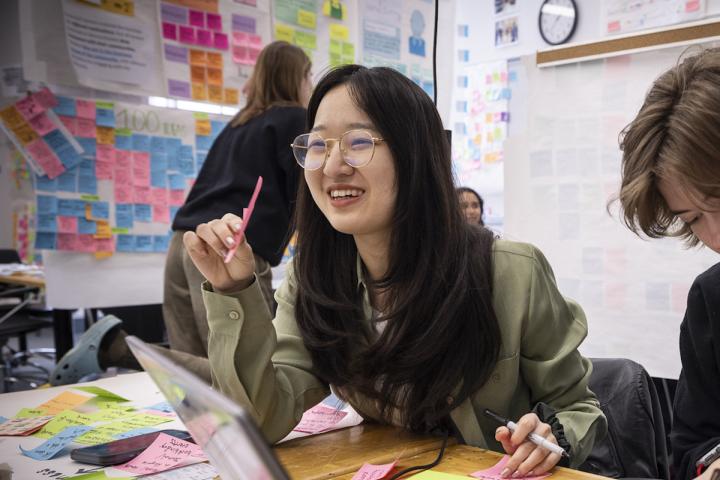 Two Olin College of Engineering students study together, with papers and bright-colored sticky notes laid out on a brown, wooded table, in one of Olin's Collaborative Design studio on campus.
